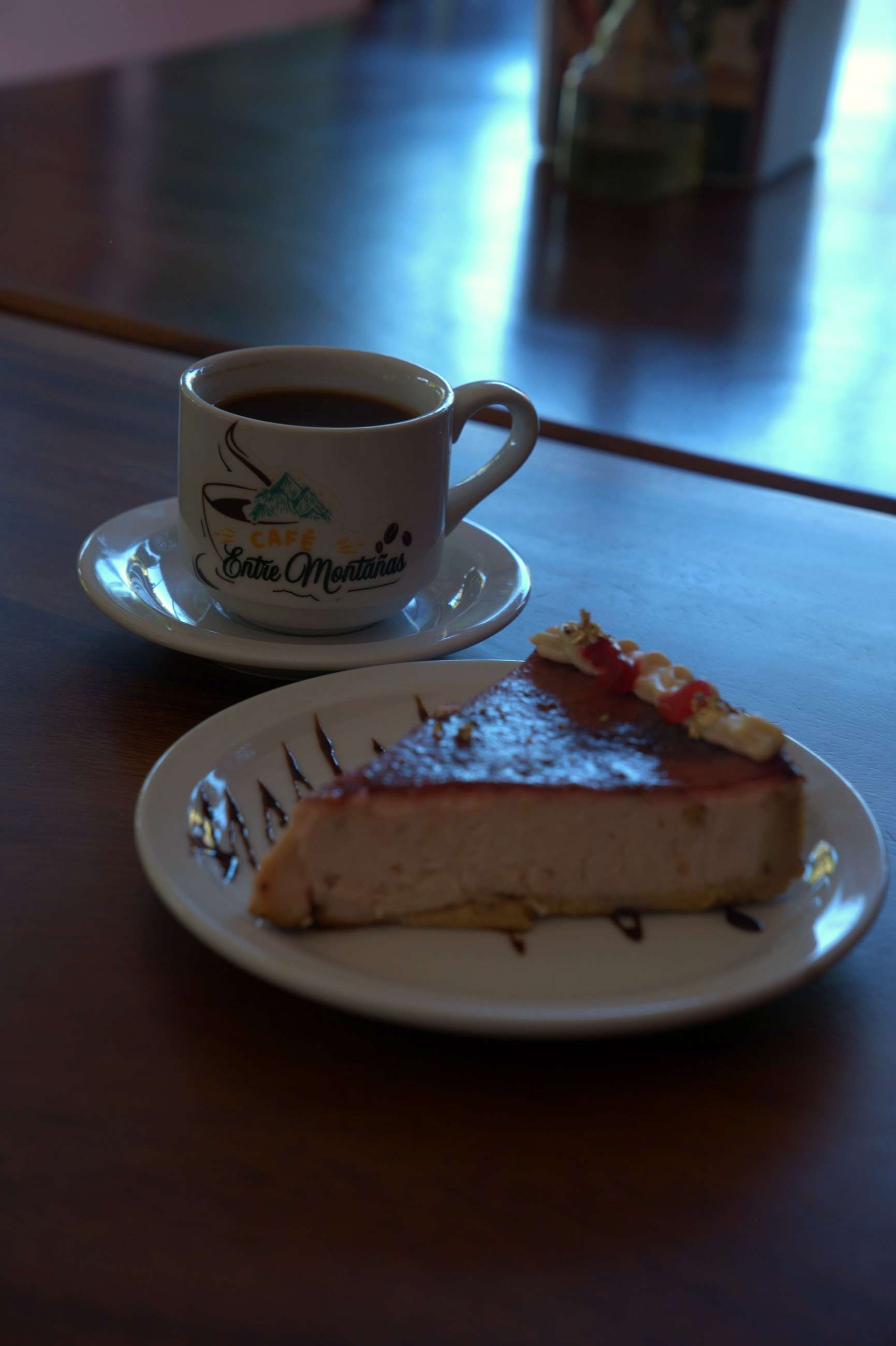 Taza de café con el logo de Café entre montañas junto a una rebanada de cheesecake